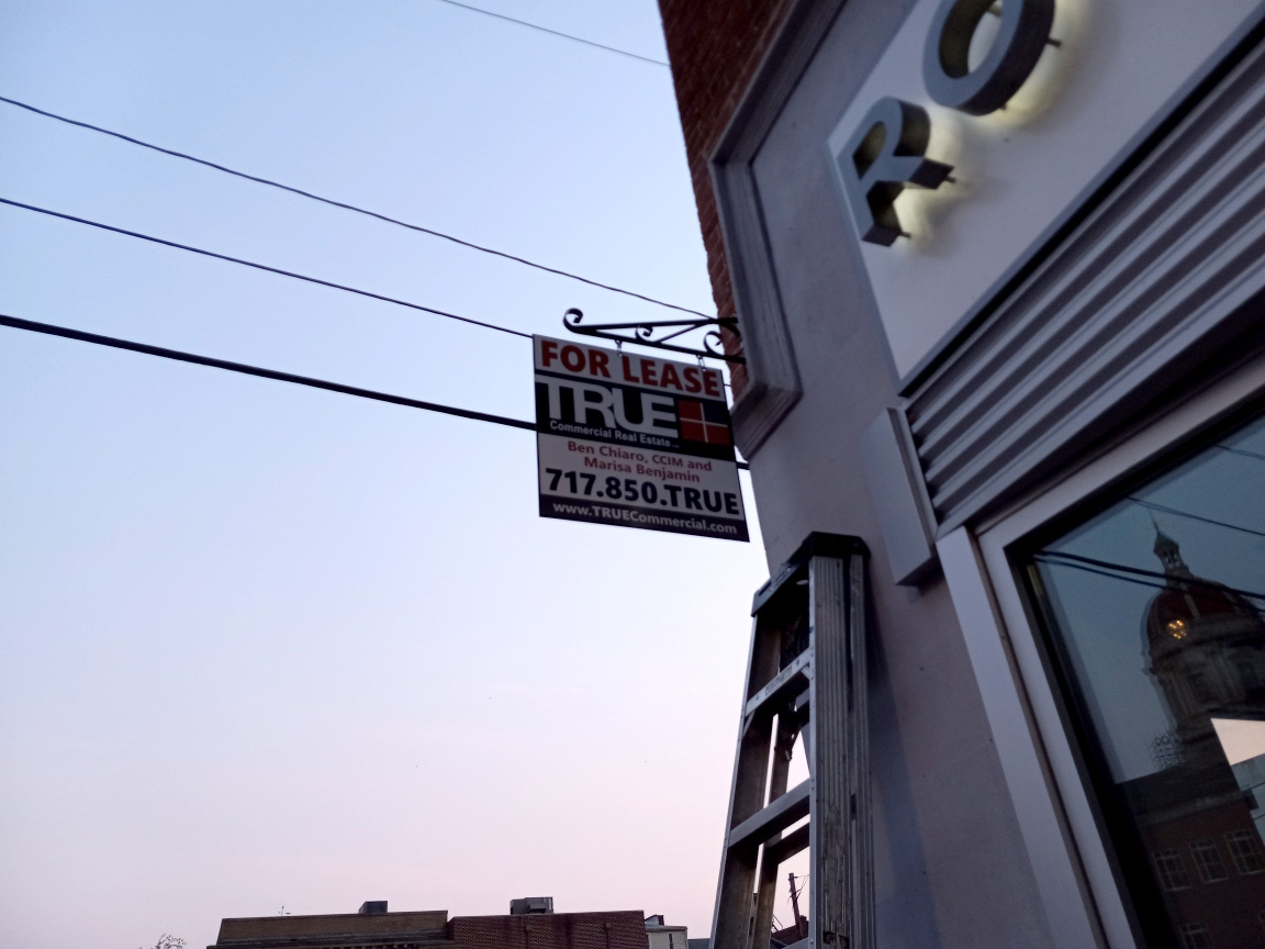 A TRUE Commercial Real Estate For Lease blade sign mounted on a decorative bracket off a downtown storefront, reading the leasing agents' names and phone number against the evening sky.